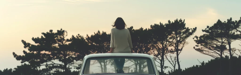 Ett fotografi i panorama-format: En ung kvinna sitter ovanpå sin bil och tittar ut över skogen och funderar över ägandekostnader.