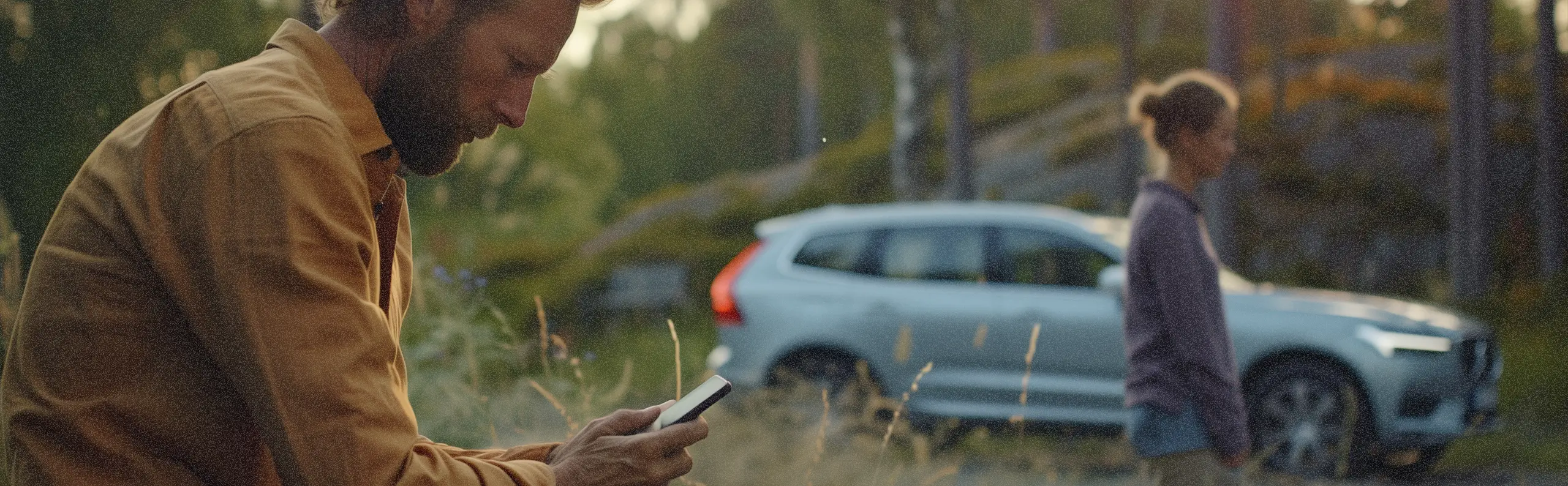 En man sitter och kollar i mobilen efter att ha krockat bilen, i bakgrunden en kvinna och en bil (volvo) i ett svenskt landskap