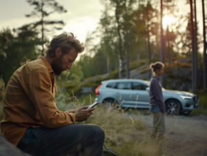 En man sitter och kollar i mobilen efter att ha krockat bilen, i bakgrunden en kvinna och en bil (volvo) i ett svenskt landskap