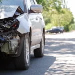 En silvrig bil som har fått en vagnskada på främre delen av bilen. Bilen står på en asfalterad vägg omringad av skog.