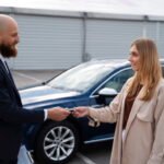 En man i mörk kostym räcker över en bilnyckel till en kvinna i en beige trenchcoat. De står på en parkeringsplats framför en mörkblå SUV, och mannen håller en blå mapp i sin andra hand.